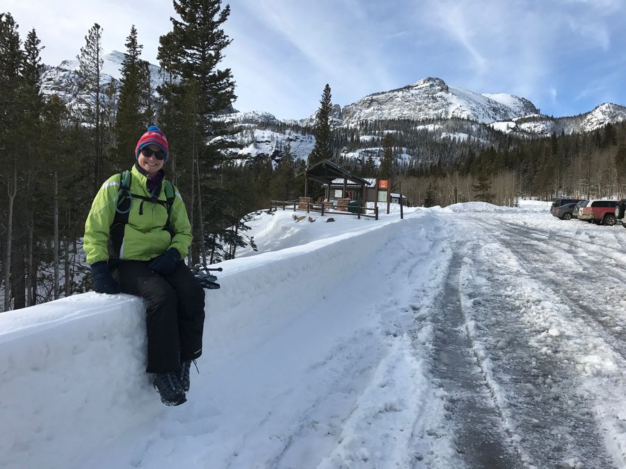 A smiling woman in a bright jacket sitting on a snowbank below snowy mountains