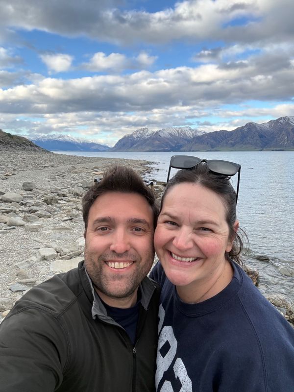 A couple taking a selfie by a lake with snowy mountains behind